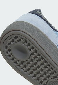 Semelle d'une basket Adidas bleu clair avec motif de grip texturé et indentation circulaire, affichant le logo de la marque sur le côté.