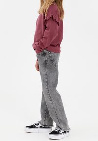 Bordeauxroter Pullover mit Rüschenärmeln, kombiniert mit grauen Jeans im geraden Schnitt und fransigem Saum, ergänzt durch schwarz-weiße Turnschuhe.