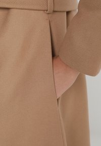 Látka hnědého vlněného kabátu s kapsou, která ukazuje hladkou texturu a ušitý design. Detailní záběr na rukáv a boční šev.