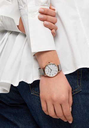 Sølvur med en tekstureret grå urskive, gulddetaljer og en sølv mesh-rem, båret på en hånd mod en hvid skjorte og blå jeans.