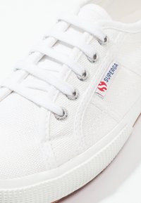 Sneaker bianco in tela con superficie testurizzata, punta rotonda, occhielli in metallo e lacci bianchi. Presenta un piccolo etichetta di marca rossa e blu sul lato.