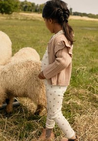 Ein Kind trägt eine beige gestrickte Strickjacke mit Rüschen über einem weißen Hemd und gemusterten Leggings. Es steht neben flauschigen Schafen auf einer grünen Wiese.