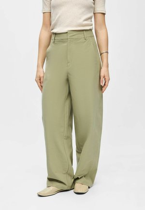 Žena oblečená v olivovo zelených širokých nohaviciach s vysokým pásom a bočnými vreckami, béžovom tričku s krátkym rukávom, béžových špicatých topánkach a striebornom náramku.
