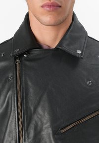 Jachetă din piele neagră, cu un finisaj texturat, guler cu capse și fermoar frontal cu accesorii din bronz. Include un buzunar lateral cu fermoar.