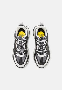 Svartvita sportskor med meshpaneler, tjock gummisula och framträdande vita snören; har en gul logotyp på innersulan.