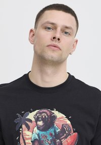 Svart bomull T-skjorte med et fargerikt grafisk trykk av en ape med solbriller på stranden, omgitt av palmer og et surfebrettdesign.