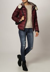 Doudoune bourgogne avec col en fourrure grise, portée sur un haut beige, associée à un jean bleu déchiré et des bottines noires.
