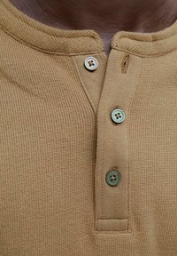 Primer plano de un cuello de camiseta de punto waffle marrón con tres botones, dos abrochados y uno desabrochado, sobre piel oscura.