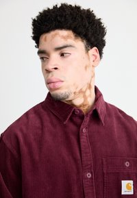 Bordeaux corduroy skjorte med krave, frontlomme og knaplukning. Har vertikal tekstur og mærke.