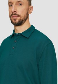 Ciemnozielona koszulka polo z długim rękawem, z klasycznym kołnierzykiem, trzema guzikami z przodu i teksturowanym wykończeniem materiału.