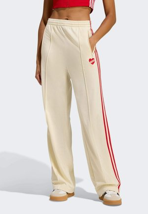 Vrouw draagt crèmekleurige broek met wijde pijpen, rode strepen en hart-logo, gecombineerd met witte sneakers en een rood cropped topje.