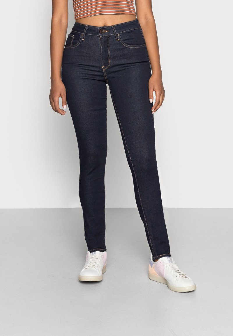 Jean taille haute bleu foncé à coupe ajustée, avec des coutures orange, des poches avant et une braguette zippée. Porté avec des baskets blanches et roses.