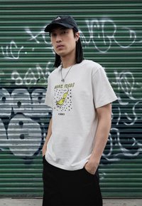 Tricou alb din bumbac cu un imprimeu grafic al unei păsări galbene și inscripția "BREAK FREE!" în verde. Modelul poartă pantaloni negri și o șapcă neagră.