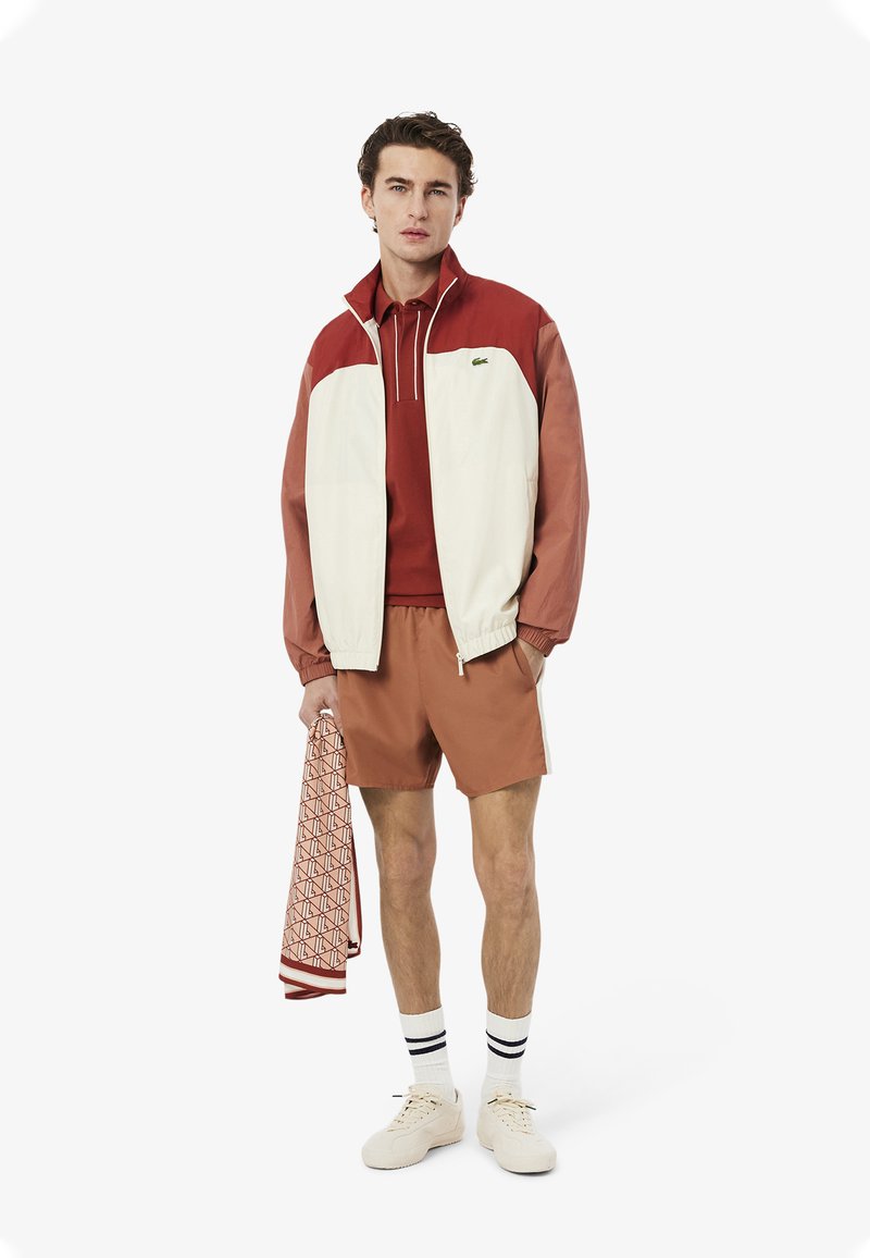 Conjunto masculino que incluye una chaqueta a rayas en blanco y rojo, polo rojo, pantalones cortos marrones, zapatillas blancas y una bufanda estampada.
