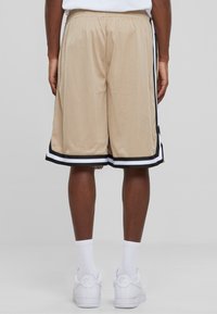 Shorts de basketball en maille beige avec des rayures noires et blanches le long de l'ourlet, taille élastique et coupe ample, vus de dos.