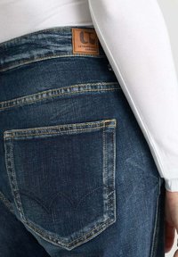 Jean en denim bleu foncé avec des coutures contrastées, présentant une étiquette en cuir marron et un design de poche distinctif. Texture douce et coupe ajustée.