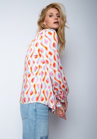 Bunte Bluse mit geometrischem Muster in Orange, Pink und Türkis, langen Ärmeln und einem leicht offenen Saum. Kombiniert mit Jeans.
