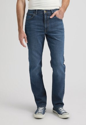 Mees, kes kannab tumedat sinist sirgete säärtega pükse, valgeid tenniseid ja valget särki, üks käsi taskus ja teine lõdvestunult kõrval.