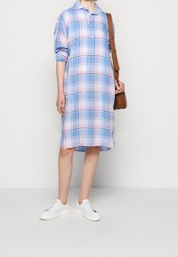 Blå och rosa rutmönstrad skjortklänning med knappfront, uppkavlade ärmar och avslappnad passform, parat med vita sneakers och en brun väska.