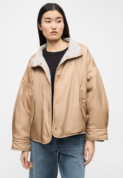 Γυναίκα που φοράει υπερμεγέθη μπεζ μπουφάν με ρίγες στο γιακά πάνω από μαύρο πουκάμισο και μπλε τζιν, στέκεται μπροστά σε απλό φόντο.