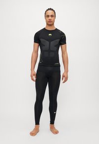 Camisa de compresión negra con diseño texturizado de músculos en el frente, combinada con leggings negros que presentan un pequeño logo blanco en la parte inferior de la pierna.