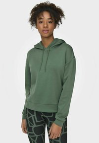 Grüner Pullover-Hoodie mit Kordelzügen, lockerer Passform, weichem Material, gerippten Bündchen und Saum. Kombiniert mit schwarzen gemusterten Leggings.