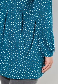 Blauwe blouse met lange mouwen en witte onregelmatige stippen, geplooide details onder de taille, gedragen met grijze pantalon.