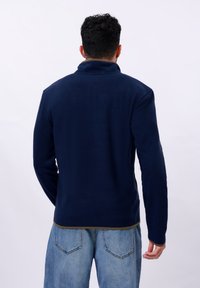 Veste en polaire marine avec un col haut, des manches longues et des bords contrastants en vert olive à l'ourlet. Texture douce, design décontracté.