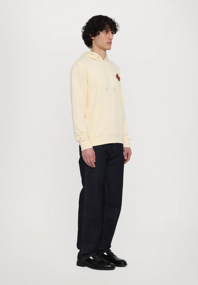 Light yellow hoodie with drawstrings, featuring a small brown logo, paired with dark blue straight-leg jeans and black shoes.