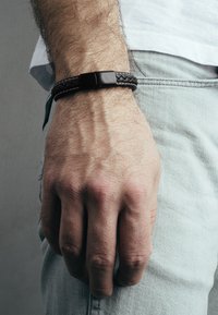 Brățară din piele împletită neagră cu o cataramă din metal negru neted, purtată pe o mână care se sprijină de o țesătură din denim de culoare deschisă. Prezintă un design texturat.