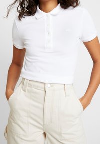 Chemise polo blanche avec un col, dotée d'une patte de boutonnage et d'un petit logo. Portée avec un pantalon beige taille haute.