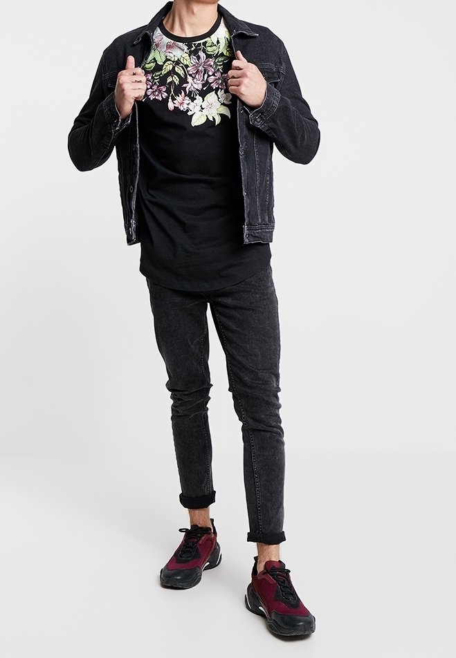 Man som bär en svart blommig t-shirt, svart denimjacka, smala svarta jeans och vinröda och svarta sneakers stående mot vit bakgrund.