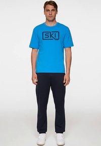 Sininen puuvillainen t-paita, jossa on lyhyet hihat ja musta suorakulmainen logo, jossa lukee "SKI." Yhdistetty tummiin housuihin ja valkoisiin kenkiin.