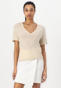Beige, rövid ujjú, V-nyakú póló könnyű anyagból, egy fehér, aszimmetrikus kérdéses szoknyával párosítva. Egyszerű, letisztult design.