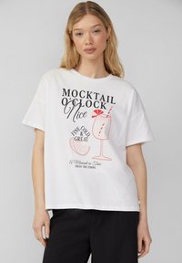T-shirt en coton blanc avec un imprimé graphique d'un verre à cocktail et un texte en noir et rouge en gras, avec une coupe décontractée et des manches courtes.
