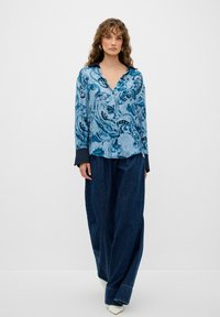 Camicia azzurro chiaro con motivo paisley, colletto e polsini neri, abbinata a pantaloni in denim scuro di taglio comodo. Tessuto liscio, vestibilità rilassata.