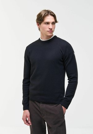 Junger Mann, der einen dunkelblauen Pullover und dunkle Hosen trägt, steht mit einer Hand in der Tasche und schaut leicht zur Seite vor einem einfarbigen Hintergrund.