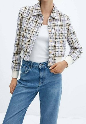 Femme portant une veste courte à carreaux avec des boutons dorés sur un débardeur blanc et un jean bleu taille haute sur un fond uni.
