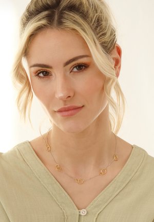 Silberne Halskette mit verbundenen goldenen Kreisen; getragen über einer hellgrünen Bluse mit strukturiertem Finish und Knopfdetails.