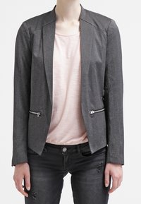 Blazer grigio con un design strutturato, caratterizzato da revers affilati, tasche frontali con zip e una texture liscia, indossato sopra un top rosa chiaro.