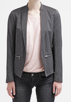 Blazer gris au design structuré, avec des revers accentués, des poches avant zippées et une texture lisse, porté sur un top rose pâle.