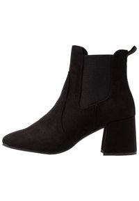 Bota corta de ante negro con puntera puntiaguda, paneles laterales elásticos y un tacón grueso, que presenta una textura suave y un diseño minimalista.