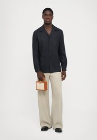 Námornícky košeľa s golierom a prednou vreckom, béžové široké nohavice, v ruke malé hnedé a béžové kabelky s hornou rúčkou.