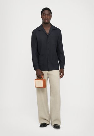 Navy skjorte med krave og lomme foran, beige brede bukser, holder en lille brune og beige håndtaske med et håndtag ovenfor.