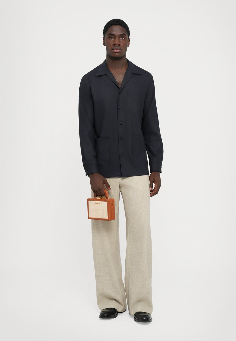 Tengerészkék galléros ing, elülső zsebbel, bézs, bő szárú nadrággal, egy kicsi barna és bézs táskával a kézben.