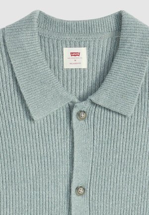 Jasnoniebieska dzianinowa koszulka polo z kołnierzykiem, pięcioma guzikami oraz metką wskazującą rozmiar medium i luźny fason. Tkanina o fakturze prążkowanej.