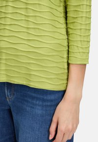 Grünes Oberteil mit texturiertem, welligem Muster, drei Viertelärmel und abgerundetem Saum, kombiniert mit blauen Jeans. Hand auf der Hüfte.