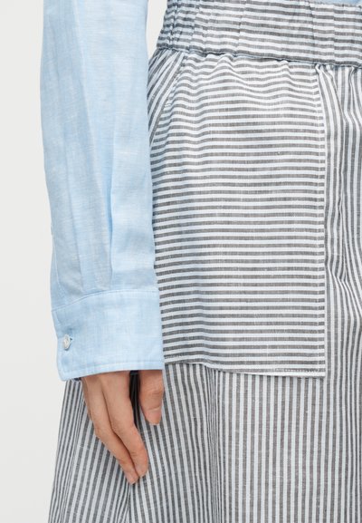 Χέρι που στηρίζεται δίπλα στη μέση, φοράει γαλάζιο πουκάμισο με μακριά μανίκια και γκρι ριγέ παντελόνια με τσέπη που ταιριάζει.