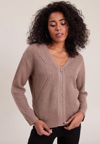 Beige gestrickte Strickjacke mit einem Reißverschluss, V-Ausschnitt und diagonalen Rippmustern. Lange Ärmel haben eine strukturierte Oberfläche.