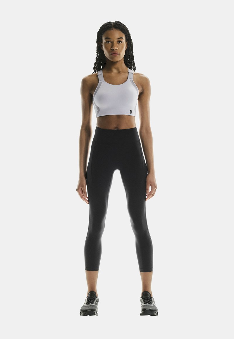 Weißer Sport-BH mit verstellbaren Trägern kombiniert mit hochgeschnittenen schwarzen Leggings. Das Model steht in schwarzen Sportschuhen. Einfaches, funktionales Design.